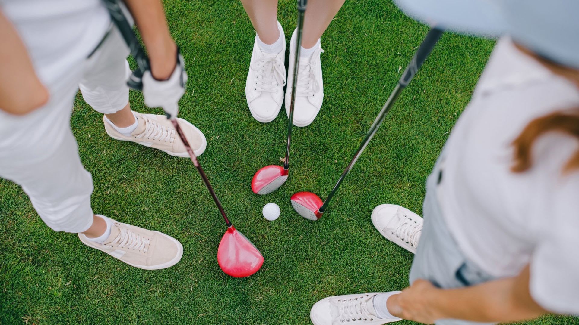 Four people wearing white sneakers from top golf brands stand on grass with golf clubs and a golf ball in the center.